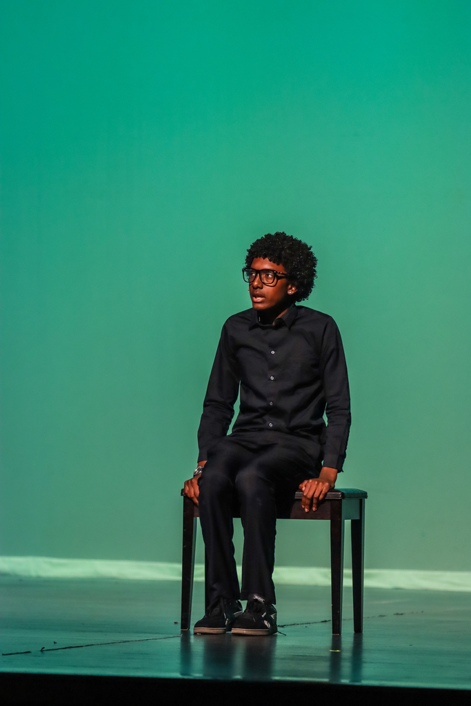 A student performer wearing glasses and an all-black outfit sits on a chair under green stage lighting during a theatrical monologue.