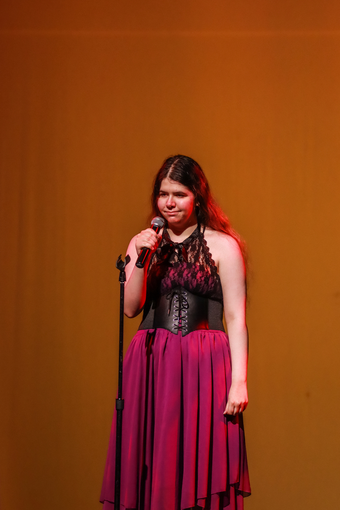 A student performer in a black and magenta formal dress stands at a microphone onstage under warm amber lighting.