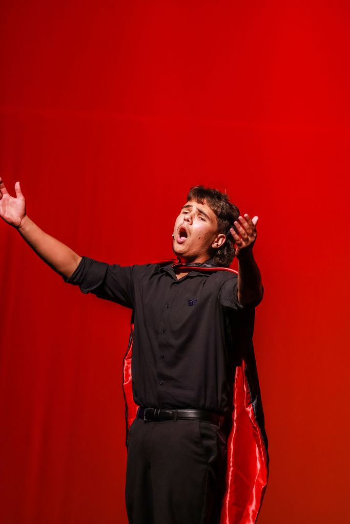 A student performer in a black outfit and red cape sings dramatically with arms outstretched against a bright red stage backdrop.