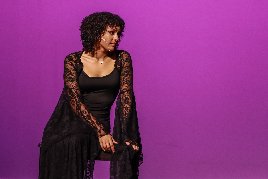 A student performer in a black dress with lace sleeves sits on a chair under purple stage lighting during a dramatic theater performance.