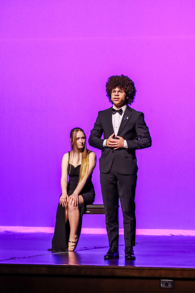 A student in a black tuxedo stands center stage speaking during a theater performance while another student in a long black dress sits behind them on a chair. Purple stage lighting fills the background, creating a dramatic atmosphere.