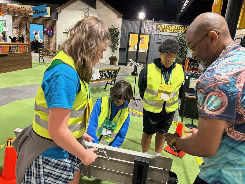 Three students wearing reflective safety vests work together with an instructor during a hands-on activity in a simulated distribution and delivery learning environment. The group is gathered around equipment while receiving guidance, with a mock town and workplace setting visible in the background.