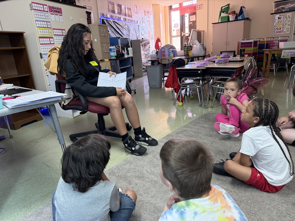 An older student sits in a classroom chair reading a booklet to a small group of elementary students seated on a rug. The younger children sit in a semicircle and focus on the reader as she shares the story. Classroom desks, learning materials, and student supplies surround the group, illustrating a peer-reading activity that encourages literacy, mentoring, and positive connections between students of different grade levels.