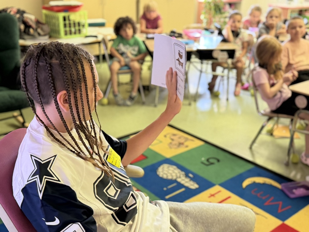 Close-up view of an older student with braided hair reading from an illustrated booklet to a classroom of younger students. The reader sits on a chair beside a colorful educational rug while elementary students watch and listen from desks in the background. The image captures student leadership and engagement during a reading lesson.