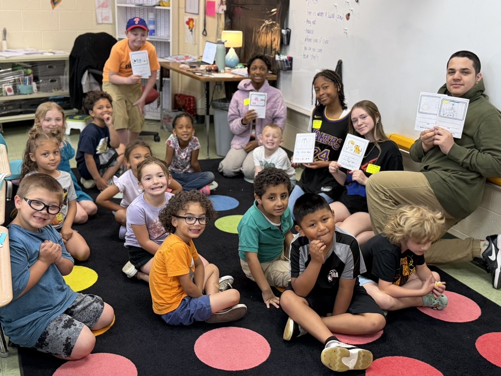 A group of elementary and secondary students gathers on a colorful classroom rug during a shared reading activity. Several older students sit and kneel among younger children while holding up illustrated booklets or student-created books. The students smile and look toward the camera, creating a welcoming atmosphere. A whiteboard, classroom shelves, and learning materials are visible in the background, highlighting a collaborative literacy experience between older and younger students.