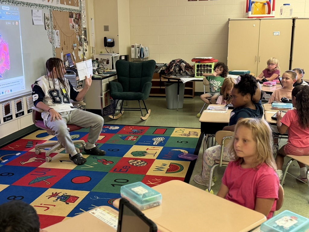 An older student sits at the front of an elementary classroom reading aloud from a booklet while younger students listen from their desks. The classroom includes a colorful alphabet-and-number rug, interactive display board, and learning centers. Students face the reader attentively, demonstrating a cross-age mentoring and literacy activity focused on reading and comprehension.
