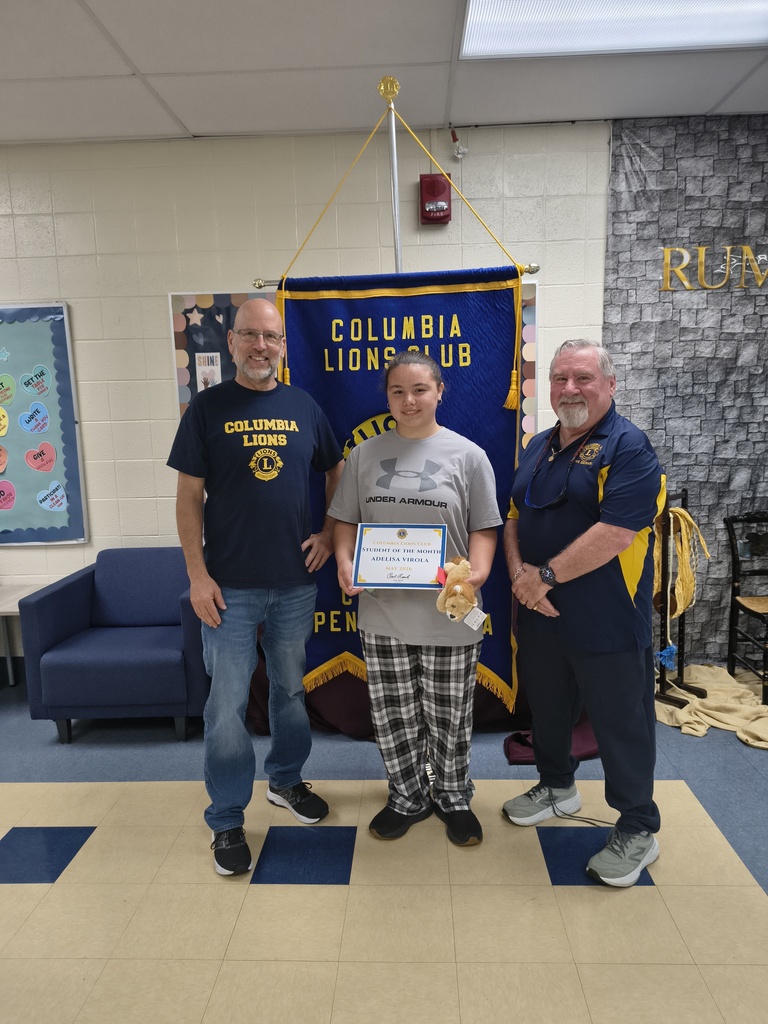 A student stands between two members of the Columbia Lions Club in front of a blue Lions Club banner. The student is holding a Student of the Month certificate and a small stuffed lion. The recognition was awarded for demonstrating the character trait of problem-solving. The photo was taken at Columbia Middle School - Taylor Campus.