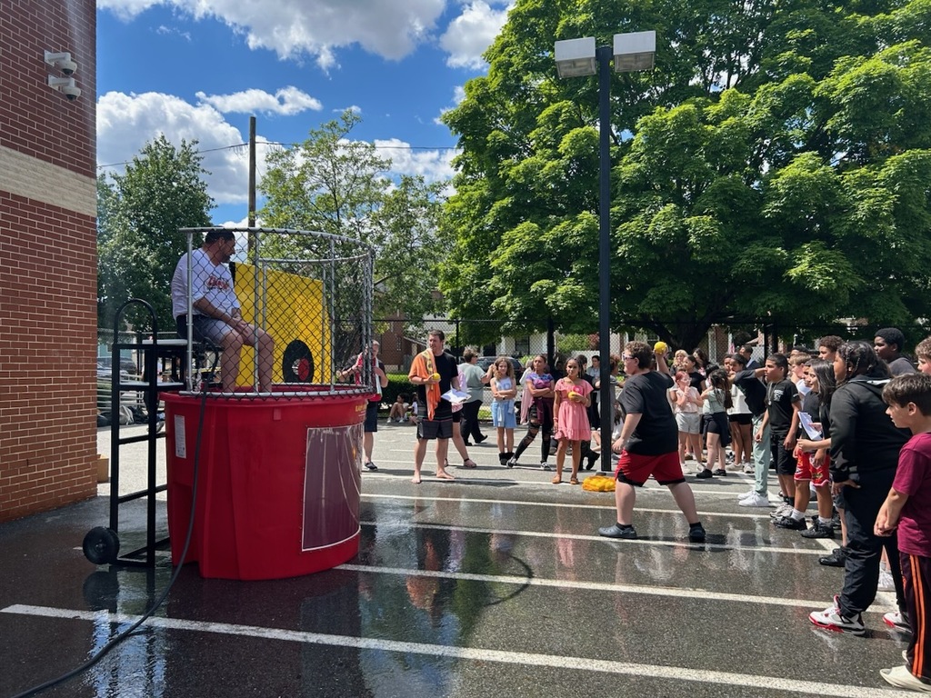 Students and staff gather around a dunk tank during an outdoor school event. A student prepares to throw a ball at the target while a staff member sits above the water tank, waiting to be dunked. The wet pavement and crowd create an energetic, fun atmosphere.