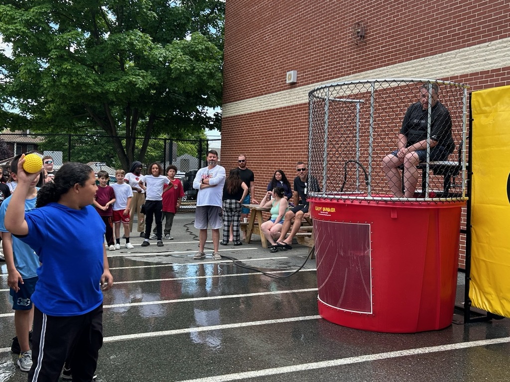 Students and staff gather around a dunk tank during an outdoor school event. A student prepares to throw a ball at the target while a staff member sits above the water tank, waiting to be dunked. The wet pavement and crowd create an energetic, fun atmosphere.
