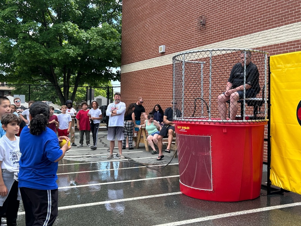 Students and staff gather around a dunk tank during an outdoor school event. A student prepares to throw a ball at the target while a staff member sits above the water tank, waiting to be dunked. The wet pavement and crowd create an energetic, fun atmosphere.