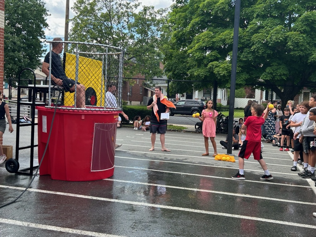 Students and staff gather around a dunk tank during an outdoor school event. A student prepares to throw a ball at the target while a staff member sits above the water tank, waiting to be dunked. The wet pavement and crowd create an energetic, fun atmosphere.