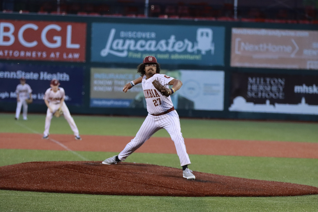 A baseball pitcher wearing a white pinstriped Columbia uniform with the number 27 stands on the mound in the middle of a pitch during a game. Another Columbia infielder is visible in the background, ready for the play, with outfield advertising signs and stadium seating behind them. The pitcher is captured with one leg extended and throwing arm drawn back, emphasizing the motion of the delivery.