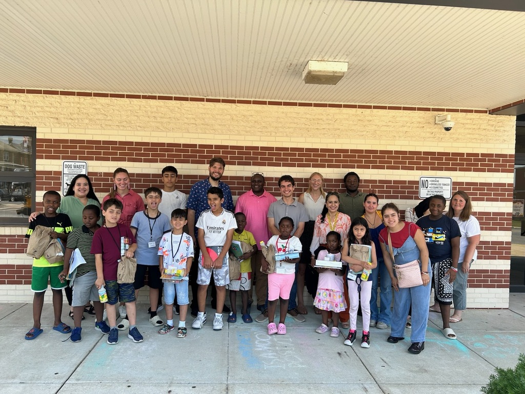 A large group of children and adults pose for a group photo outside a brick building entrance under a covered walkway. The group includes approximately 15 children and 10 adults standing together and smiling at the camera. Several children are holding brown paper bags, notebooks, and small items. The adults stand behind the children, creating a supportive community atmosphere. The photo appears to be taken during a summer youth program or educational camp gathering on a sunny day.
