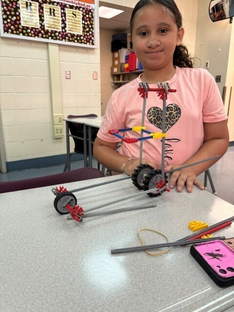 A student stands proudly beside a STEM engineering project built from K'NEX pieces on a classroom table. The structure features gears, rods, wheels, and a tall frame connected by rubber bands and support beams. The student is wearing a light pink shirt with a heart design and smiling at the camera. Additional K'NEX pieces and a smartphone are visible on the table, while classroom posters, shelves, and chairs can be seen in the background.