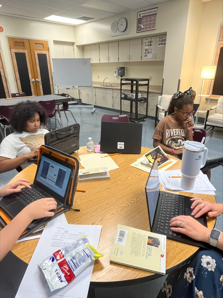 Four students and a teacher sit around a round table in a classroom during Columbia Middle School - Taylor Campus's Summer Literacy Camp. Students are reading, writing, and working on laptops while discussing activities related to their book study. Books, worksheets, pencils, and laptops are spread across the table as participants engage in collaborative literacy and research tasks. The classroom features cabinets, a whiteboard, and comfortable seating areas, creating a welcoming learning environment.