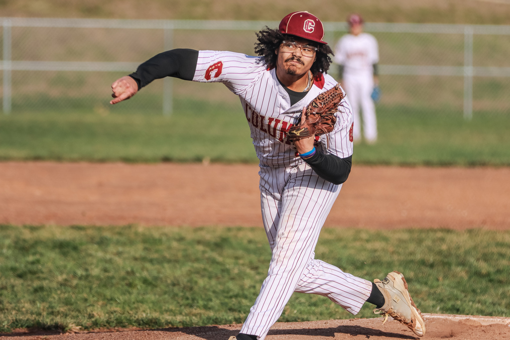 A Columbia High School baseball pitcher in a white pinstriped uniform and maroon cap throws a pitch during a game. He is captured mid-delivery on the mound with his glove raised and throwing arm extended. Another player stands blurred in the outfield, with the infield dirt, grass, and chain-link fence visible in the background.
