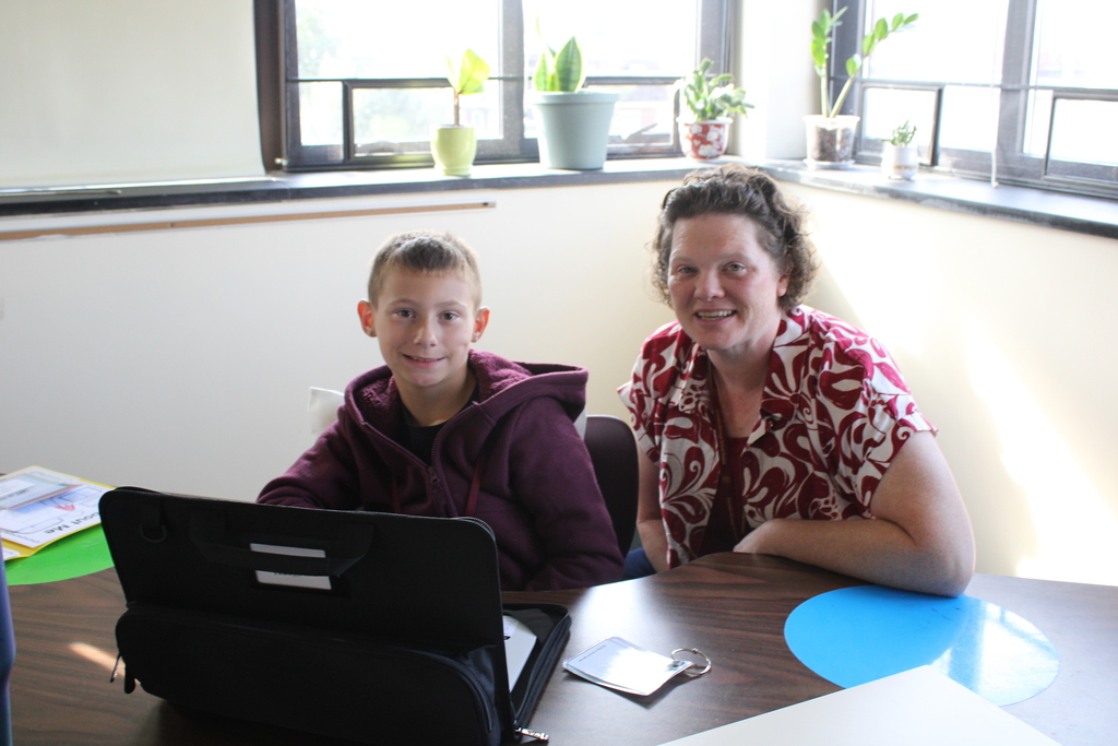 A student sits beside a teacher at a classroom table, both smiling toward the camera while working with a laptop.