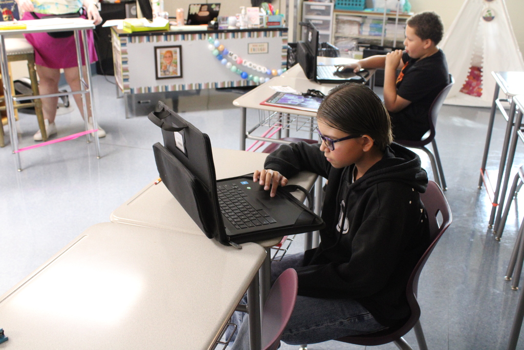 A student works on a laptop at a classroom desk while another student works at a desk in the background.