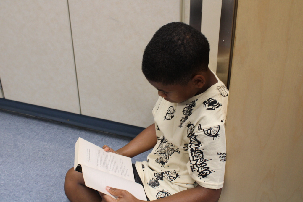Young boy seated on a hallway floor, quietly reading an open book; his graphic-print shirt and focused expression create a calm mood.
