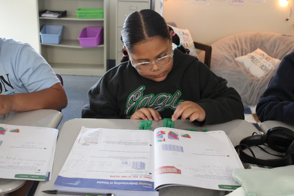 A student works on a math activity at a classroom desk, using green manipulatives alongside an open math workbook.