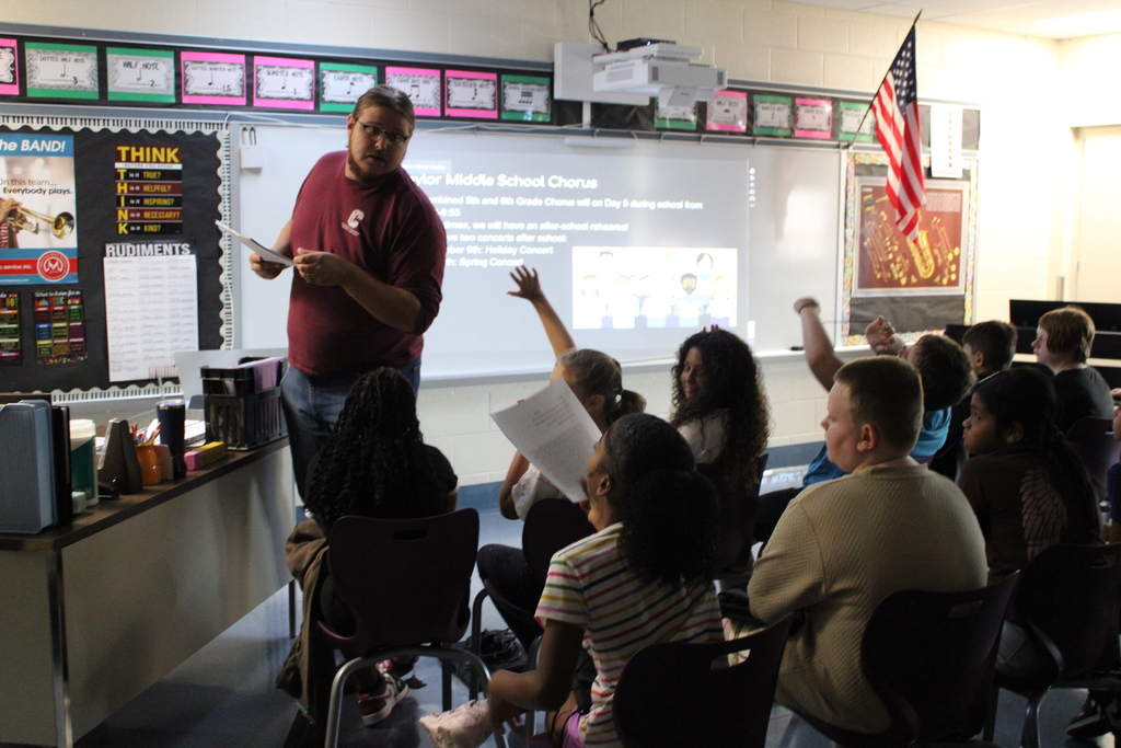 Teacher leads an interactive classroom lesson as students raise their hands, with a projected slide and American flag in the background.