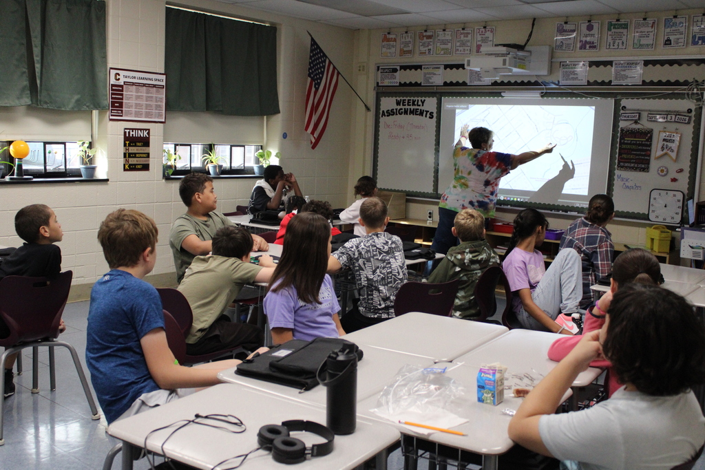 Elementary students watch an instructor point to a projected lesson in a lively classroom, surrounded by desks, posters, and an American flag.
