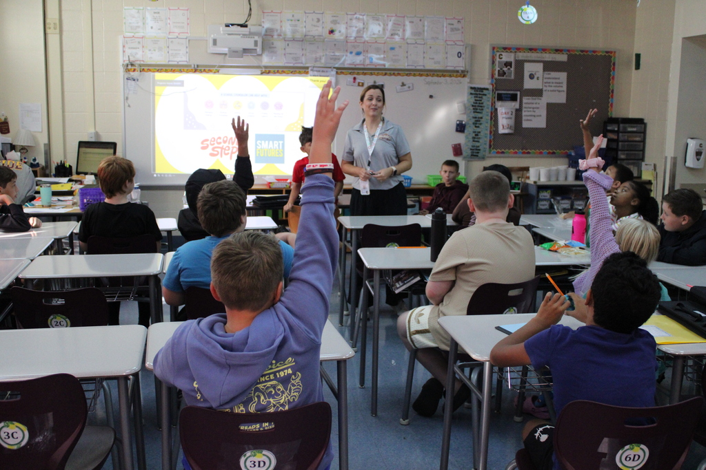 Teacher leads an engaging classroom lesson as several students raise their hands, with desks, a projected screen, and colorful posters visible.