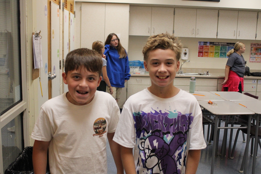 Two smiling boys pose in a classroom, one wearing a white T-shirt and the other a purple graphic shirt; students and desks fill the background.