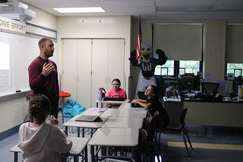 A teacher speaks to students in a classroom as a costumed eagle mascot raises its wings near an American flag, creating a playful mood.