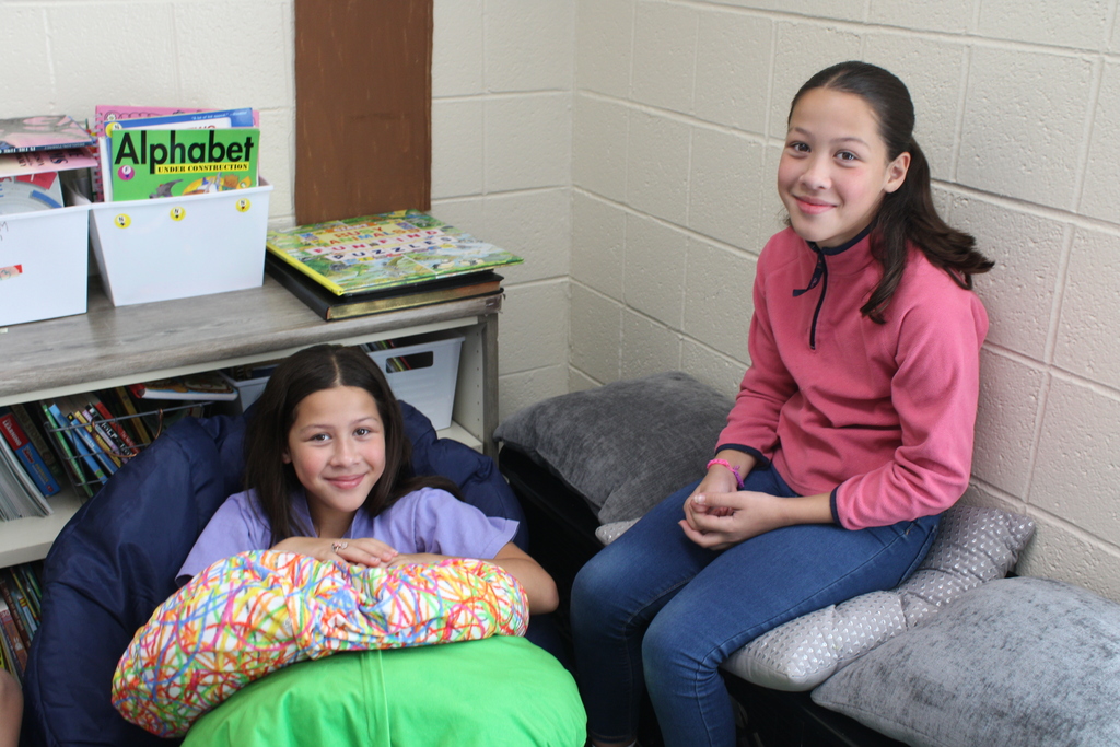 Two students smile while sitting together in a cozy classroom reading area surrounded by books, cushions, and colorful pillows.