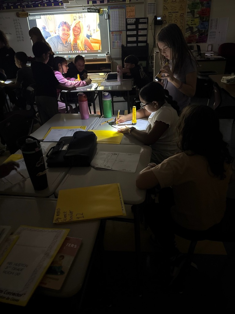 Students work together at desks in a dimly lit classroom while a large screen displays a photo. Worksheets, folders, and water bottles are scattered across the desks.