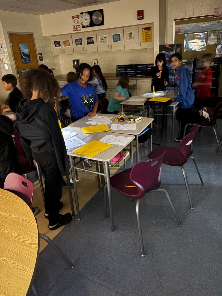 Students work and move around a brightly lit classroom, with desks grouped together and covered with worksheets, folders, books, and pencils. Posters and storage cabinets line the walls.