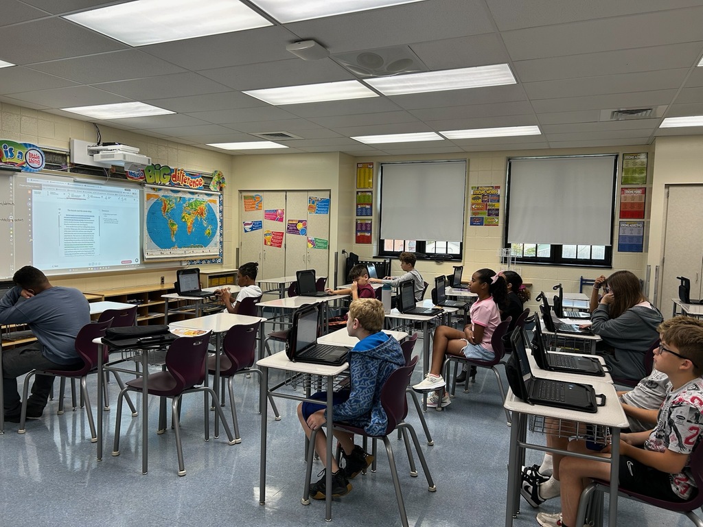 Students sit at individual desks in a middle school classroom, working on laptops and listening to instruction. A projected lesson is displayed at the front of the room beside a world map, while colorful educational posters and charts decorate the walls.