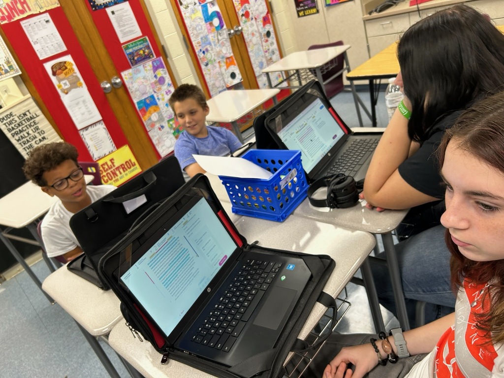 Students sit at individual desks in a middle school classroom, working on laptops and listening to instruction. A projected lesson is displayed at the front of the room beside a world map, while colorful educational posters and charts decorate the walls.