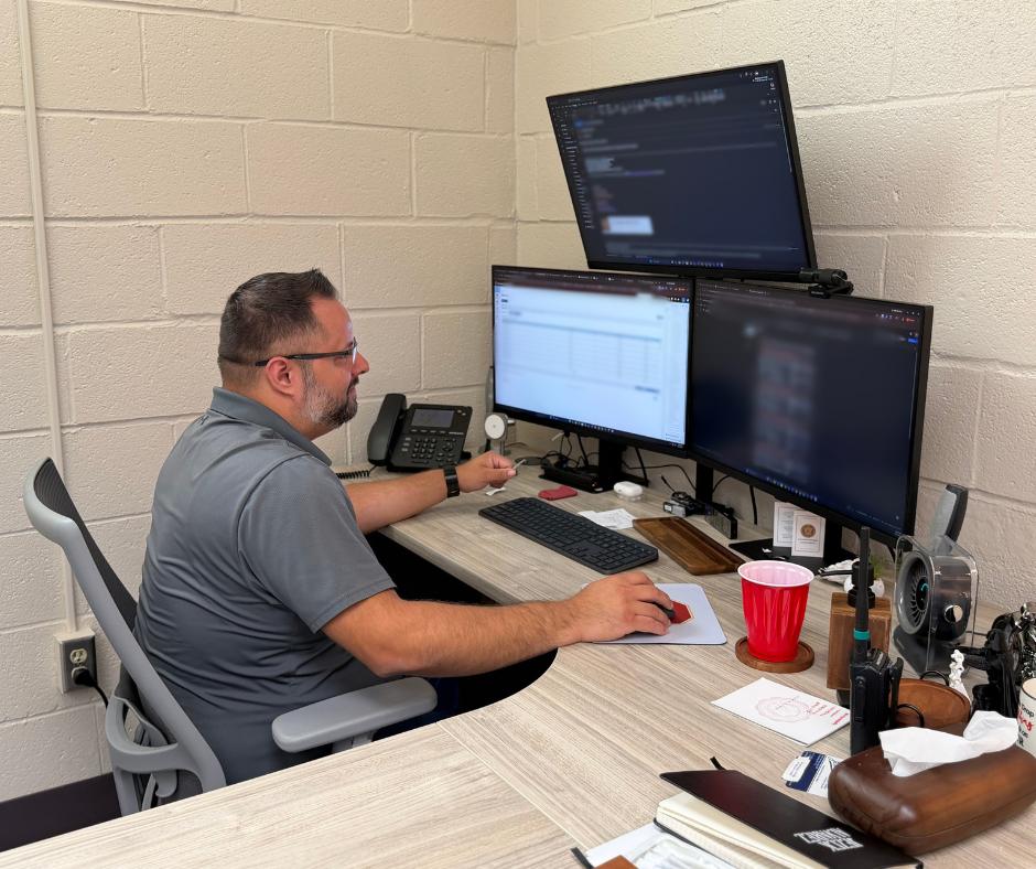 Technology staff member works at a desk with three computer monitors displaying technical information.