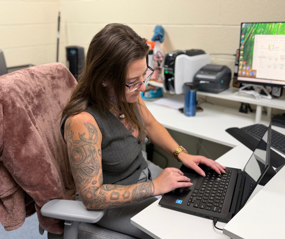 Technology staff member uses a laptop at a desk with a computer monitor, printer, keyboard, and office equipment.