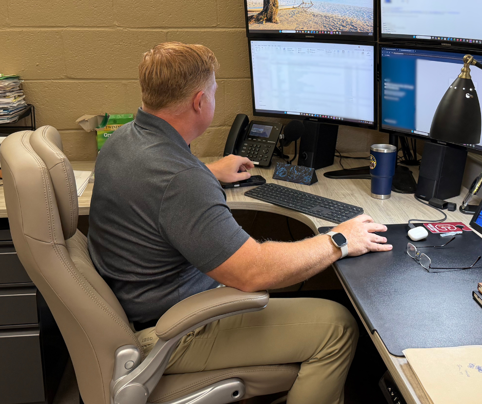 Technology staff member works at a desk with four computer monitors, a keyboard, phone, and office supplies.