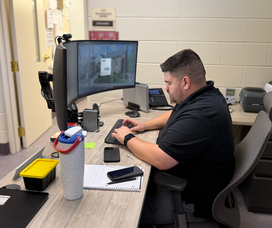 Technology staff member types at a desk with two computer monitors, a laptop, phone, notebook, and office equipment.