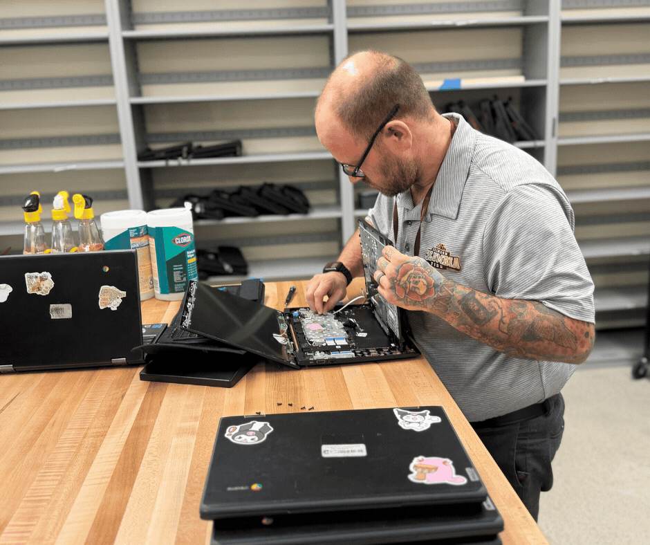image of a technology staff member repairing an open laptop on a worktable, with additional laptops and computer equipment nearby.