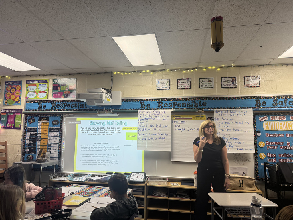 A 5th grade teacher leads a personal narrative writing lesson while students work at their desks. The classroom displays reminders about “Showing, Not Telling,” personal narratives, and using evidence in writing.