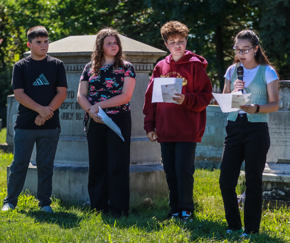 Four students stand among cemetery monuments as one student reads from a paper into a microphone.