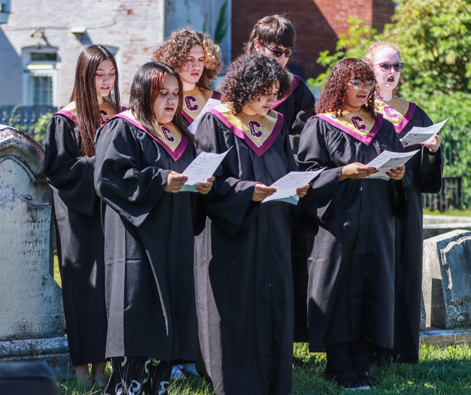 Students wearing choir gowns stand in a cemetery and sing while holding printed programs.
