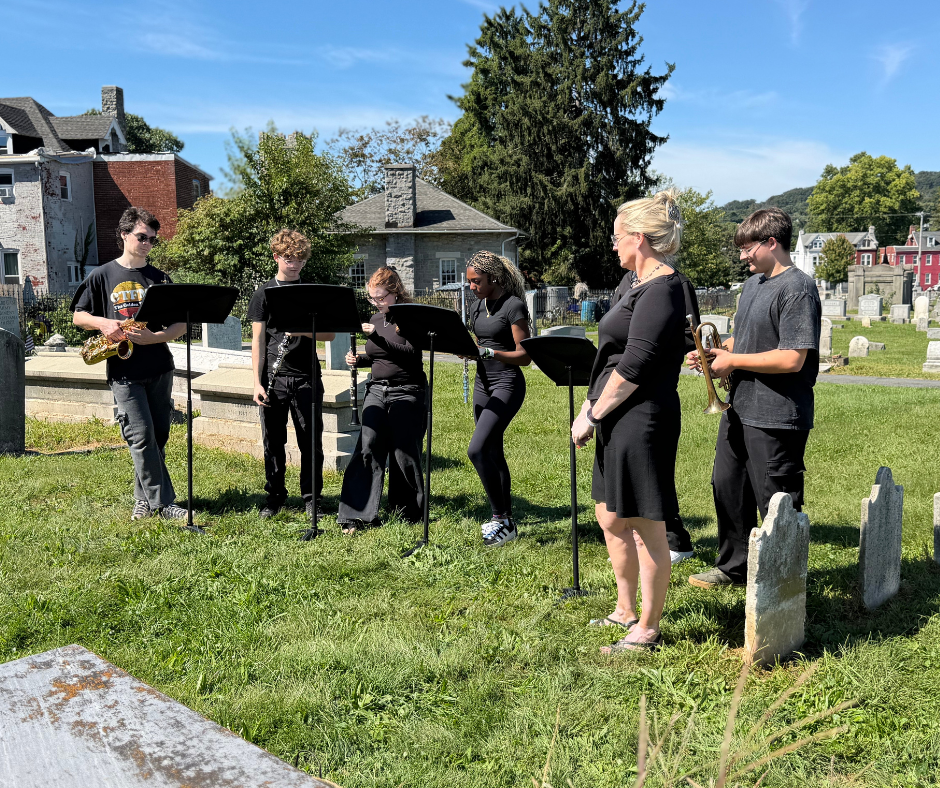 Students wearing gowns stand in a cemetery and sing while holding printed programs.