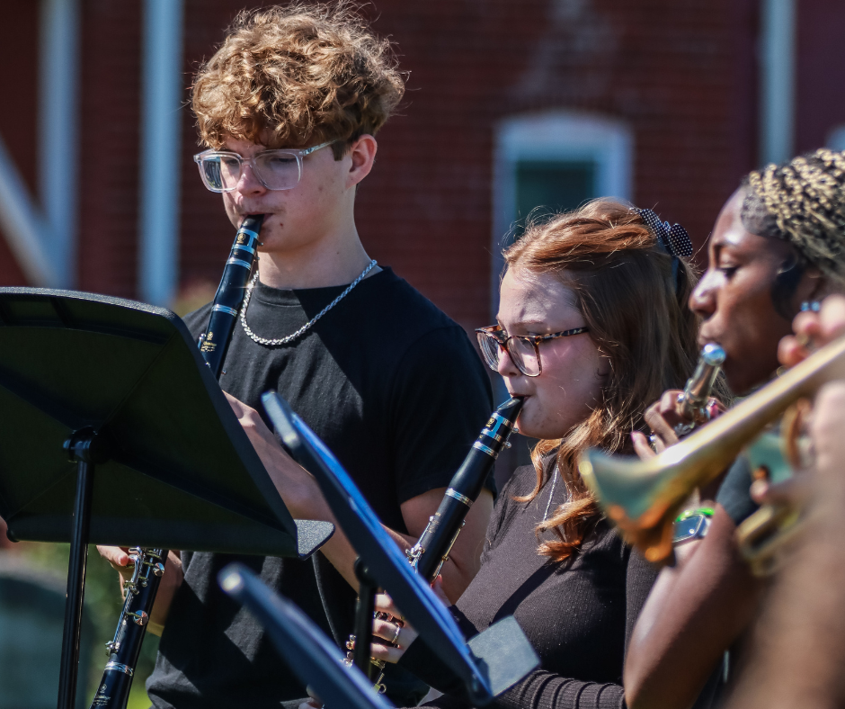 Student musicians and an adult stand among cemetery monuments during an outdoor performance, with saxophone, clarinet, and trumpet players positioned behind music stands.