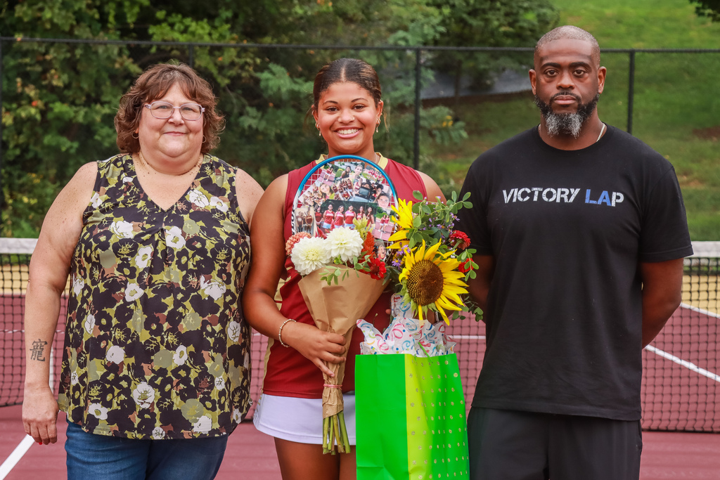 Senior Columbia tennis player stands on the court with two family members, holding flowers, a decorated tennis racket, and a green gift bag.