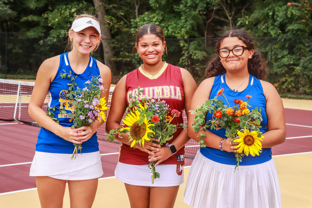 Columbia senior tennis player poses with two Northern Lebanon seniors on the tennis court. All three hold bouquets; the Columbia player wears a maroon “Columbia” uniform, while the Northern Lebanon players wear blue uniforms.