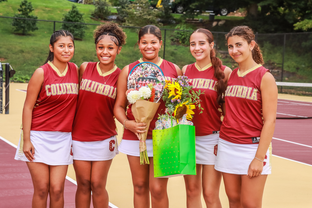 Columbia senior tennis player poses with four teammates on the tennis court. The players wear maroon Columbia uniforms, and the senior holds flowers, a decorated tennis racket, and a green gift bag.