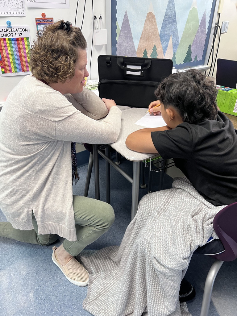 A teacher sits beside a student at a desk and supports him as he writes on a worksheet.