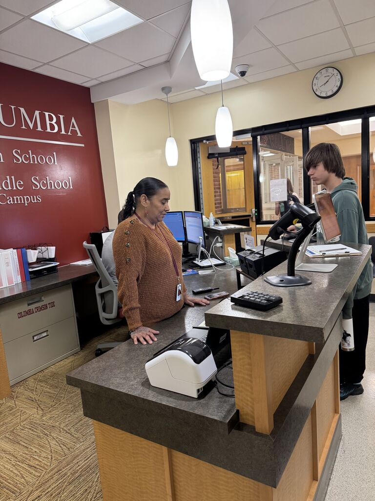 A school office staff member assists a student at the middle school reception desk.