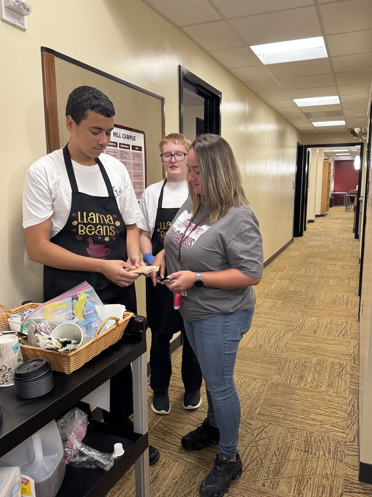 Two student baristas wearing Llama Beans aprons serve a staff member from a mobile coffee cart in a school hallway.